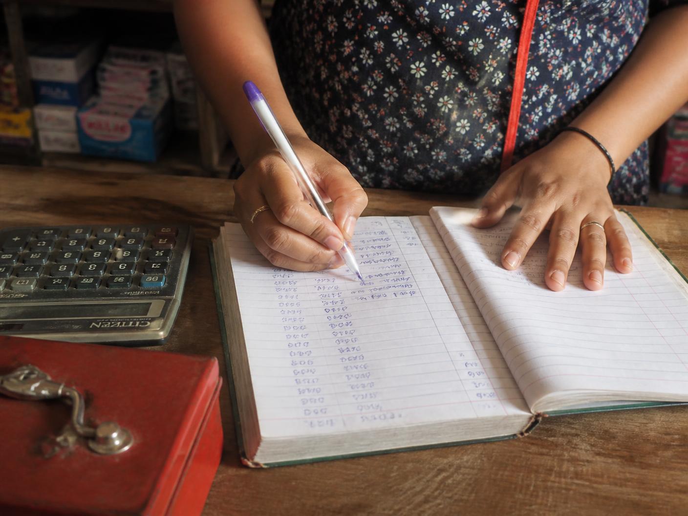 Close-up of a shopkeeper's hands writing daily sales into a paper ledger beside a calculator